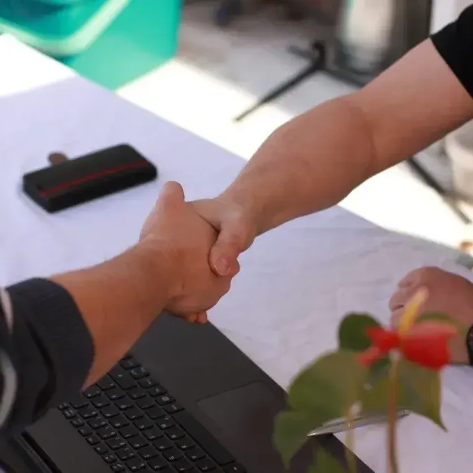 Elektriker mit fairen Preise Zwei Personen schütteln sich die Hände über einem Laptop, mit einer Blume im Vordergrund.
